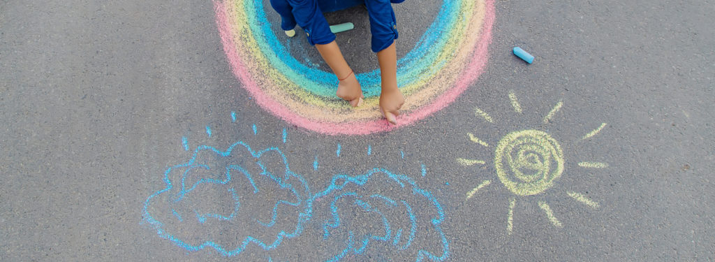 A child draws a rainbow, sun and clouds on pavement.