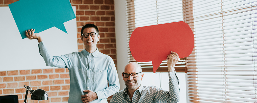 Tow men in an office setting hold cut outs of cartoon speech bubbles over their heads